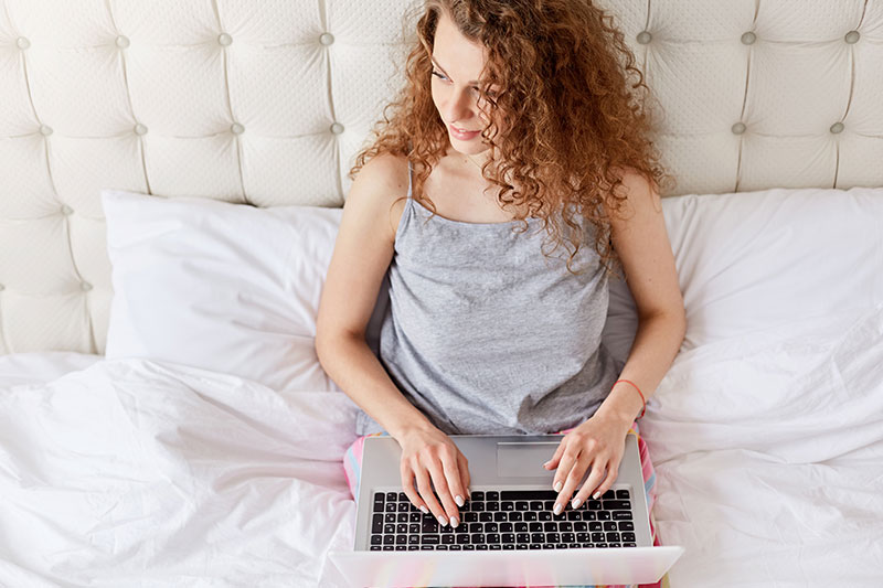 Frau mit lockigen Haaren sitzt mit Laptop im Bett