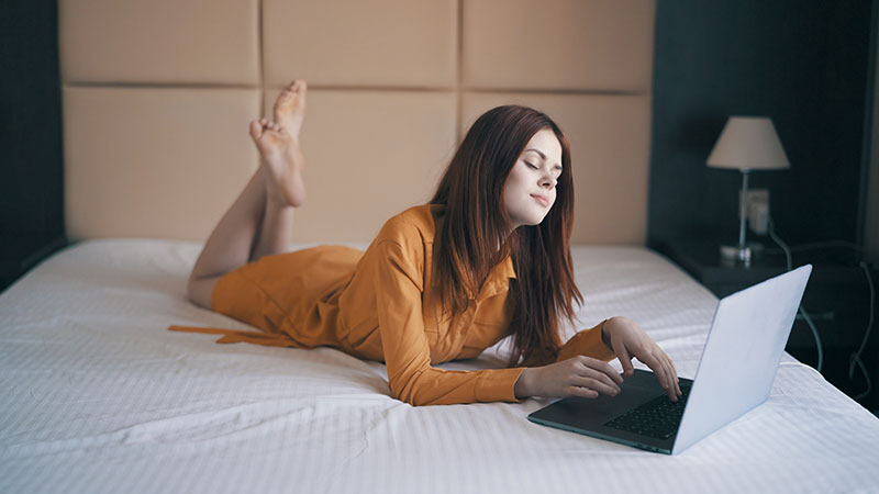 Frau liegt im Bett und tippt auf einem Laptop