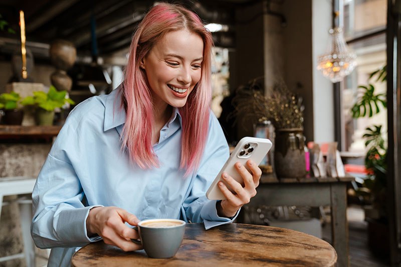 Frau mit rosa Haaren sitzt trinkt Kaffee und hat ein Handy in der Hand