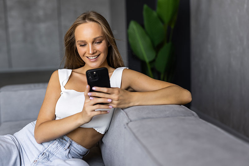 Junge Frau im Top sitzt auf Couch und tippt am Smartphone