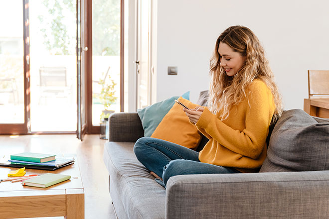 Blonde Frau sitzt auf Sofa und schaut glücklich ins Handy