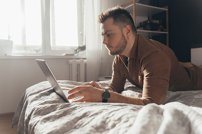 Mann mit Bart liegt auf dem Bett mit einem Laptop